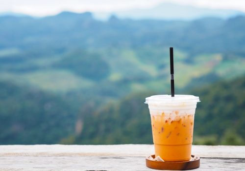 Glass of ice milk tea on vintage wooden table over green forest on the mountain background.